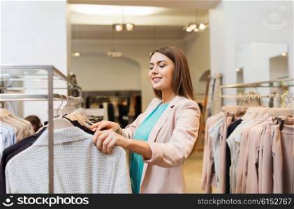 sale, shopping, fashion, style and people concept - happy young woman choosing clothes in mall or clothing store