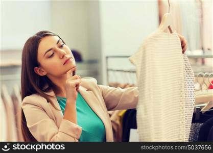 sale, shopping, fashion, style and people concept - pensive young woman choosing clothes in mall or clothing store. thoughtful young woman choosing clothes in mall