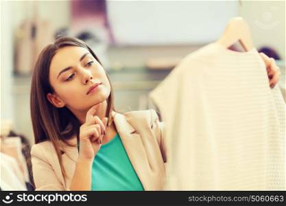 sale, shopping, fashion, style and people concept - pensive young woman choosing clothes in mall or clothing store. thoughtful young woman choosing clothes in mall