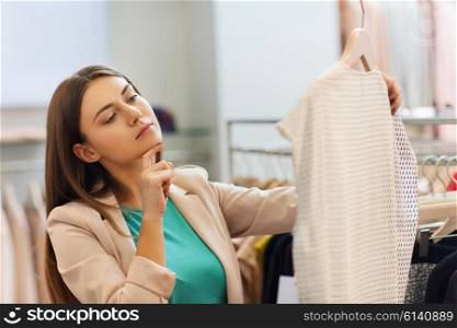 sale, shopping, fashion, style and people concept - pensive young woman choosing clothes in mall or clothing store