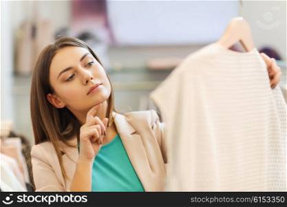 sale, shopping, fashion, style and people concept - pensive young woman choosing clothes in mall or clothing store