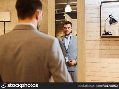 sale, shopping, fashion, style and people concept - young man choosing and trying suit on and looking to mirror in mall or clothing store
