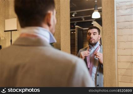 sale, shopping, fashion, style and people concept - young man choosing and tying tie on and looking to mirror in mall or clothing store