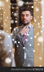 sale, shopping, fashion, style and people concept - young man choosing and trying jacket on and looking to mirror in mall or clothing store over snow