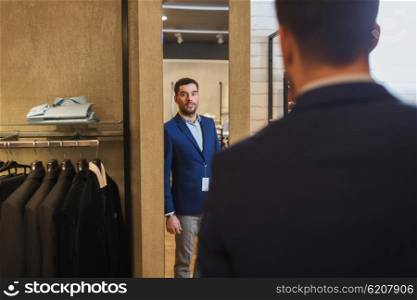 sale, shopping, fashion, style and people concept - young man choosing and trying jacket on and looking to mirror in mall or clothing store