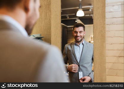 sale, shopping, fashion, style and people concept - young man choosing and trying jacket or suit on and looking to mirror in mall or clothing store