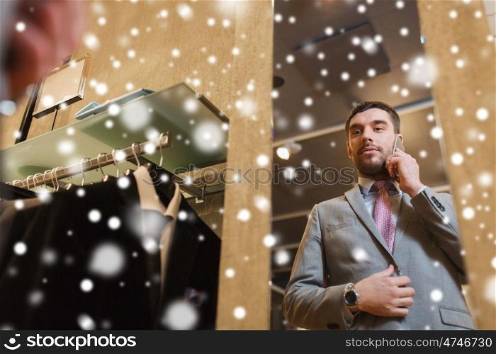 sale, shopping, fashion, style and people concept - young man or businessman trying suit and tie on and calling on smartphone at clothing store mirror over snow