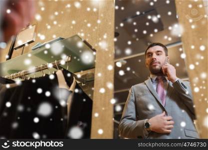 sale, shopping, fashion, style and people concept - young man or businessman trying suit and tie on and calling on smartphone at clothing store mirror over snow