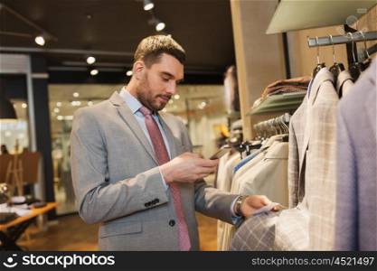sale, shopping, fashion, technology and people concept - happy man in suit with smartphone choosing clothes at clothing store