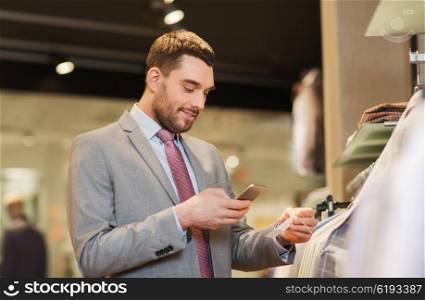 sale, shopping, fashion, technology and people concept - happy man in suit with smartphone choosing clothes at clothing store