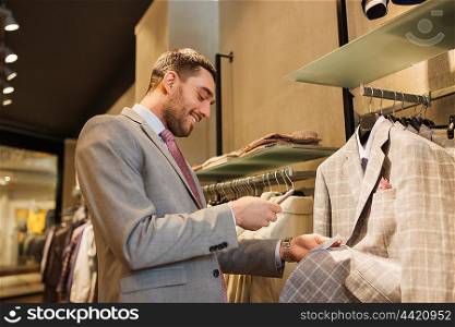 sale, shopping, fashion, technology and people concept - happy man or businessman in suit with smartphone choosing clothes at clothing store