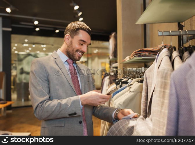 sale, shopping, fashion, technology and people concept - happy man or businessman in suit with smartphone choosing clothes at clothing store