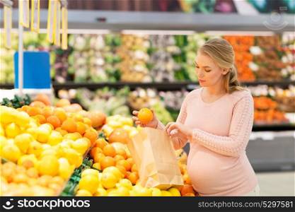 sale, shopping, food, pregnancy and people concept - happy pregnant woman buying oranges and putting them into paper bag at grocery store or supermarket. pregnant woman with bag buying oranges at grocery