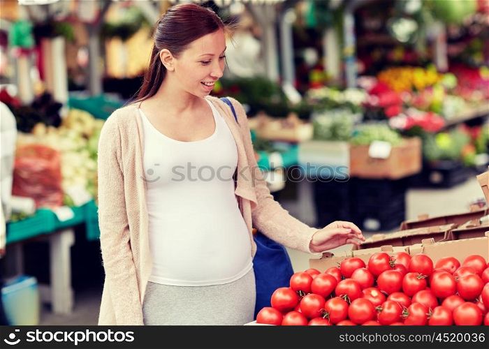 sale, shopping, food, pregnancy and people concept - happy pregnant woman choosing tomatoes at street market