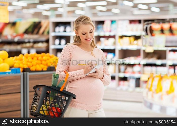 sale, shopping, food, pregnancy and people concept - happy pregnant woman with basket and notebook at grocery store or supermarket. pregnant woman with shopping basket at grocery