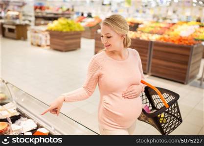 sale, shopping, pregnancy and people concept - happy pregnant woman with basket pointing finger to food on stall at grocery store or supermarket. pregnant woman with shopping basket at grocery