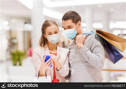 sale, technology and pandemic concept - happy young couple with shopping bags and smartphone wearing face protective medical mask for protection from virus disease in mall. couple in masks with smartphone in shopping mall