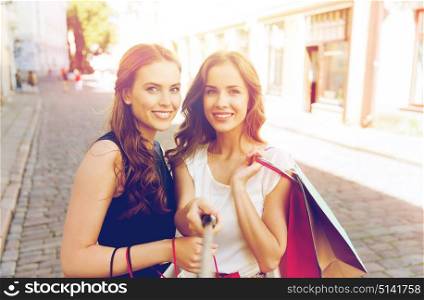 sale, technology, friendship and people concept - happy young women with shopping bags taking picture by smartphone selfie stick on city street. women shopping and taking selfie by smartphone