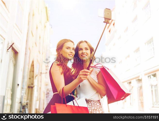 sale, technology, friendship and people concept - happy young women with shopping bags taking picture by smartphone selfie stick on city street. women shopping and taking selfie by smartphone