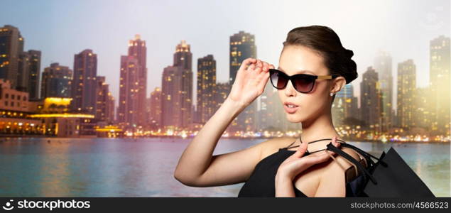 sale, tour, fashion, people and luxury concept - happy beautiful young woman in black sunglasses with shopping bags over dubai city night lights background