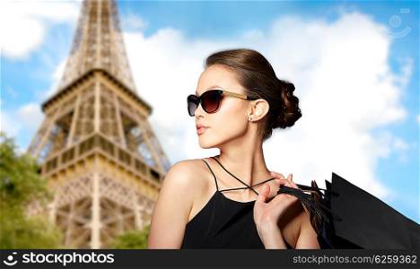 sale, tour, fashion, people and luxury concept - happy beautiful young woman in black sunglasses with shopping bags over paris eiffel tower background