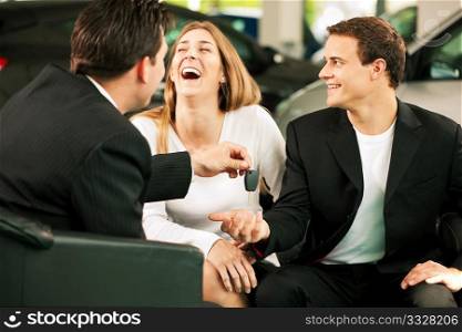 Sales situation in a car dealership, the dealer is handing auto keys to a young couple, they are excited, cars standing in the background