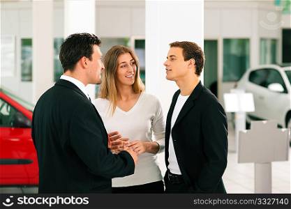 Sales situation in a car dealership, the dealer is talking to a young couple, there are cars standing in the background