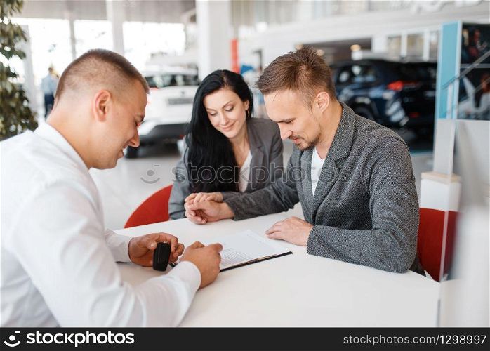 Salesman and a couple make out the sale of a new car in the showroom. Male and female customers looks vehicle in dealership, automobile sale, auto purchase