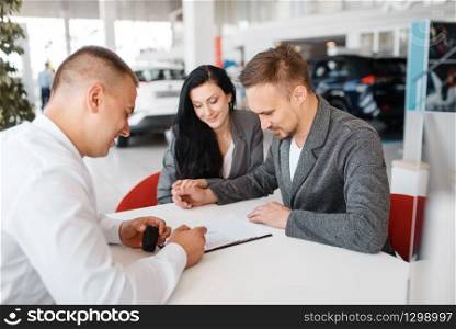 Salesman and a couple make out the sale of a new car in the showroom. Male and female customers looks vehicle in dealership, automobile sale, auto purchase