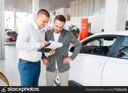 Salesman and buyer make out the purchase of new car in showroom. Male customer buying vehicle in dealership, automobile sale