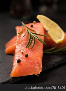 Salmon fillet with lemon and rosemary on black slate plate - close up photography of steak of fresh red fish with spices on dark background for healthy seafood and dieting concept.. Salmon fillet with lemon and rosemary on black slate plate.