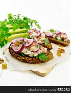 Salmon, petiole celery, raisins, walnuts, red onions and curd cheese salad on toasted bread with green lettuce leaves on parchment on a wooden board background
