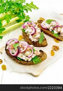 Salmon, petiole celery, raisins, walnuts, red onions and curd cheese salad on toasted bread with green lettuce on paper on a white wooden board background