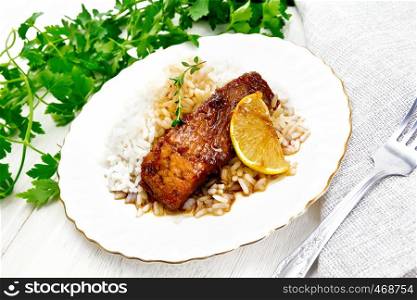 Salmon with sauce of honey, lemon juice, garlic, hot pepper and soy sauce, boiled rice, a slice of lemon and sprig of thyme in a plate, parsley, towel and fork on the background of wooden board