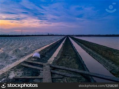 Salt farm in the morning with sunrise sky and clouds. Landscape of sea salt field in Thailand. Sea water in canal and soil pathway in farm. Raw material of salt industrial. Summer tourism in Thailand.