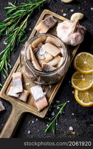 Salted herring in a jar on a cutting board with rosemary and lemon. On a black background. High quality photo. Salted herring in a jar on a cutting board with rosemary and lemon.