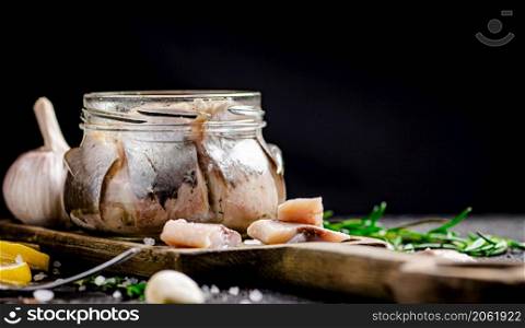 Salted herring in a jar on a cutting board with rosemary and lemon. On a black background. High quality photo. Salted herring in a jar on a cutting board with rosemary and lemon.