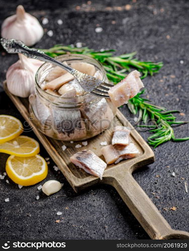 Salted herring in a jar on a cutting board with rosemary and lemon. On a black background. High quality photo. Salted herring in a jar on a cutting board with rosemary and lemon.