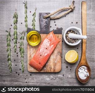 salted salmon fillet on a cutting board with rosemary, pepper, oil, lemon and a wooden spoon with salt on wooden rustic background top view