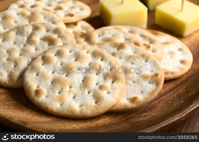 Saltine or soda crackers with cheese pieces on wooden plate, photographed with natural ight (Selective Focus, Focus in the middle of the first cracker)