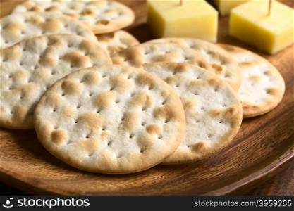 Saltine or soda crackers with cheese pieces on wooden plate, photographed with natural ight (Selective Focus, Focus in the middle of the first cracker)