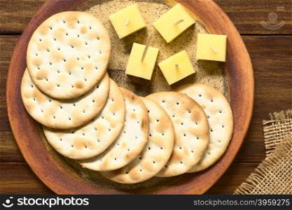 Saltine or soda crackers with cheese pieces served on plate, photographed overhead on dark wood with natural ight