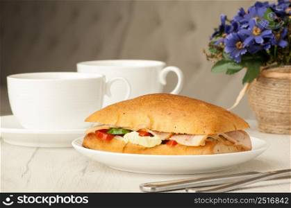 sandwich and two cups of coffee on a table with cutlery and flowers. sandwich on the plate and cups on the table