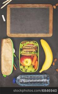 Sandwich, apple, grape, carrot, berry in plastic lunch boxes, stationery and bottle of water on black chalkboard. Back to school concept.