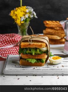 sandwich of French toast and lettuce leaves and boiled egg, a vegetarian food wrapped in paper on a white wooden board. French toast and lettuce leaves and boiled egg