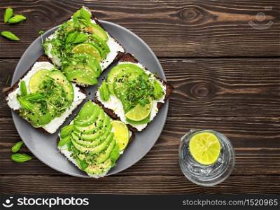 Sandwich on rye bread with avocado, cheese, microgreens, sesame seeds and spruce tips in plate on dark wooden table. Healthy vegetarian snack, flat lay with copy space. Lemonade with lime on table. Sandwich with microgreens, cheese, avocado and spruce tips. Top view with copy space, tasty and healthy vegetarian breakfast, copy space