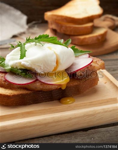 sandwich on toasted white slice of bread with poached eggs, green leaves of arugula and radish, morning breakfast on a brown wooden board
