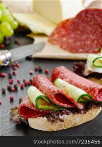Sandwich with salami, cheese, bacon, lettuce and cucumber. Quick breakfast. Black stone background. On the back side are the head of cheese, salami, spices. Close-up.
