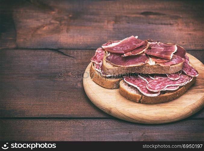 sandwiches with smoked sausage salami and pieces of hamon on a round wooden board, empty space on the left