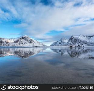 Sandy beach with beautiful reflection in water, Lofoten islands, Norway. Landscape with snowy mountains, sea, blue sky with clouds reflected in water in winter. Nature background with rocks and coast